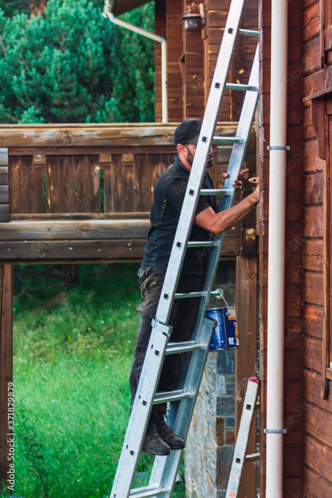 Man on ladder varnishing the wooden wall of a chalet with a terrace behind him Man on ladder varnishing the wooden wall of a chalet with a terrace behind him