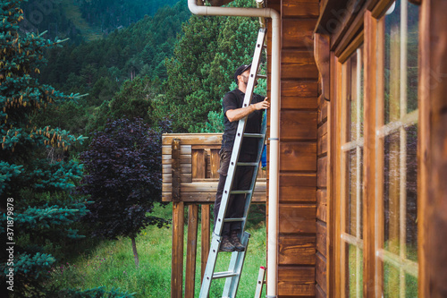 Man on a ladder working on a wooden cottage with trees and mountains behind