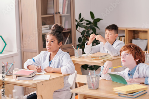 Youthful serious mixed-race girl of elementary age in whitecoat making notes