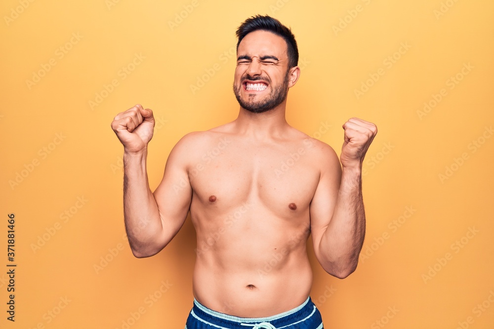 Young handsome man with beard wearing sleeveless t-shirt standing over yellow background very happy and excited doing winner gesture with arms raised, smiling and screaming for success. Celebration