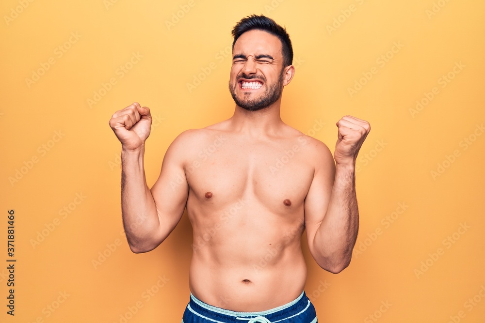 Young handsome man with beard wearing sleeveless t-shirt standing over yellow background very happy and excited doing winner gesture with arms raised, smiling and screaming for success. Celebration Young handsome man with beard wearing sleeveless t-shirt standing over yellow background very happy and excited doing winner gesture with arms raised, smiling and screaming for success. Celebration