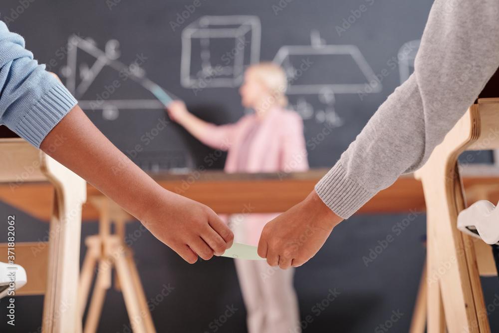 Hands of two schoolkids passing each other small notepaper with right answer