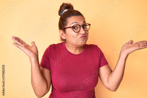 Middle age brunette hispanic woman wearing casual clothes and glasses clueless and confused with open arms, no idea and doubtful face.