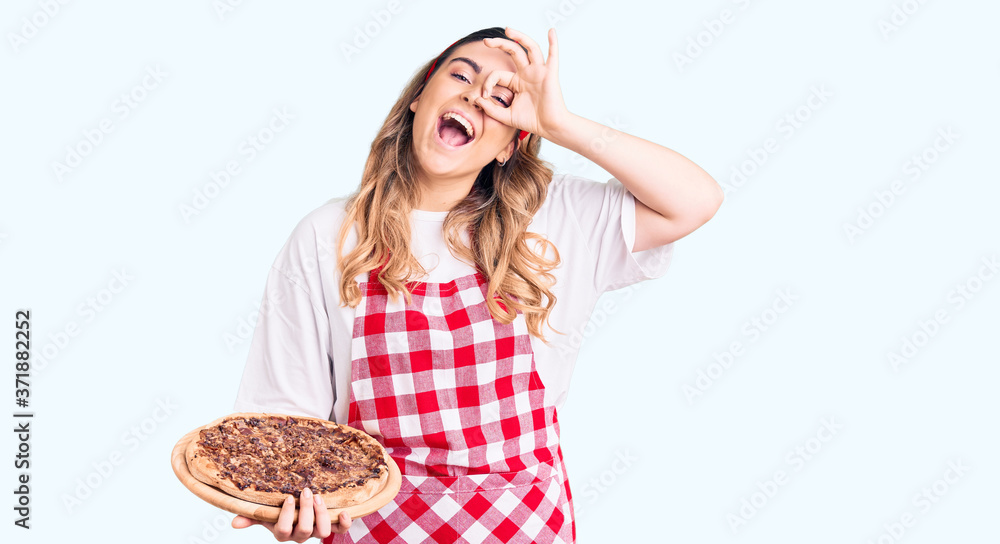 Young caucasian woman wearing apron holding pizza smiling happy doing ok sign with hand on eye looking through fingers Young caucasian woman wearing apron holding pizza smiling happy doing ok sign with hand on eye looking through fingers