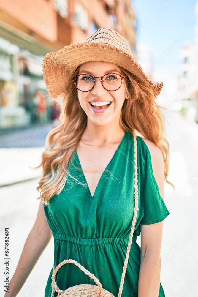 Young beautiful blonde woman on vacation wearing summer hat and glasses smiling happy. Standing with smile on face at street of city.