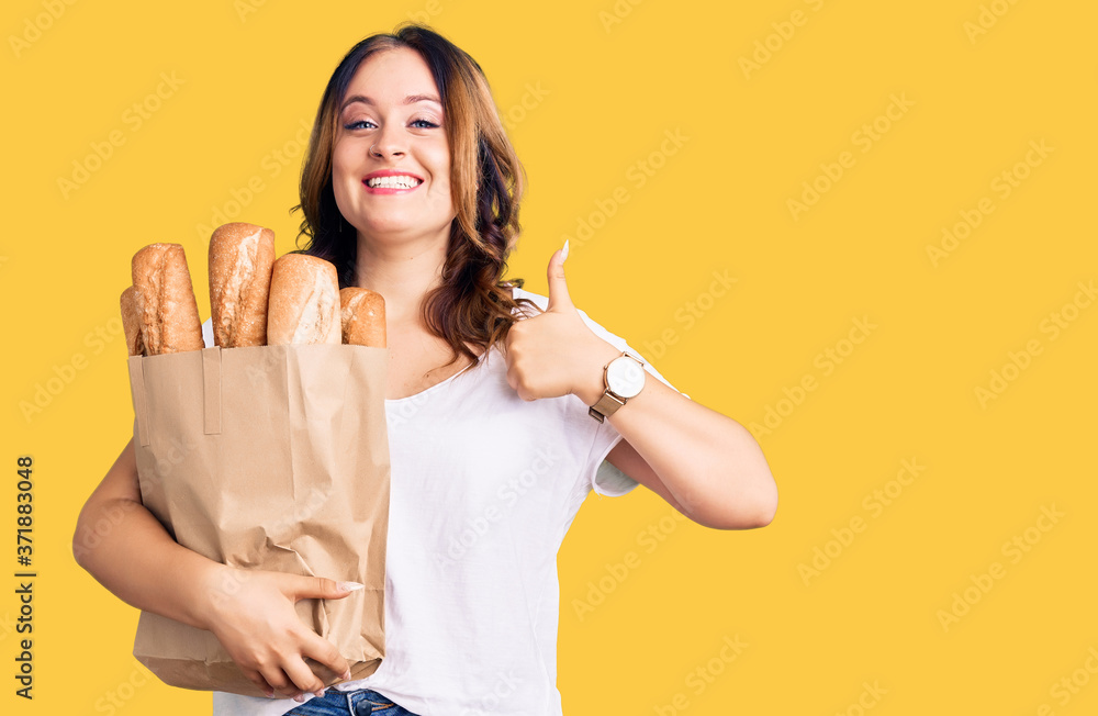 Young beautiful caucasian woman holding paper bag with bread smiling happy and positive, thumb up doing excellent and approval sign Young beautiful caucasian woman holding paper bag with bread smiling happy and positive, thumb up doing excellent and approval sign