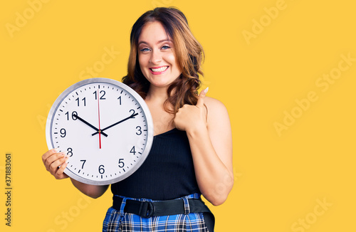 Young beautiful caucasian woman holding big clock smiling happy and positive, thumb up doing excellent and approval sign