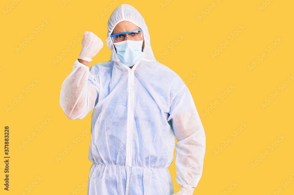 Young hispanic man wearing doctor protection coronavirus uniform and medical mask angry and mad raising fist frustrated and furious while shouting with anger. rage and aggressive concept. Young hispanic man wearing doctor protection coronavirus uniform and medical mask angry and mad raising fist frustrated and furious while shouting with anger. rage and aggressive concept.