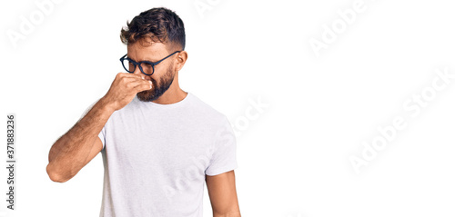 Фотография Young hispanic man wearing casual clothes and glasses smelling something stinky and disgusting, intolerable smell, holding breath with fingers on nose