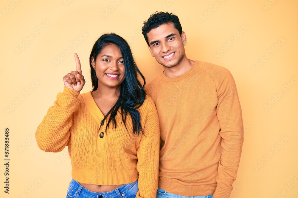 Fototapeta premium Beautiful latin young couple wearing casual clothes together showing and pointing up with finger number one while smiling confident and happy.