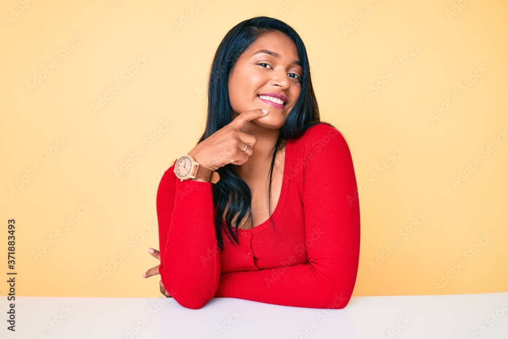 Beautiful latin young woman with long hair wearing casual clothes sitting on the table smiling looking confident at the camera with crossed arms and hand on chin. thinking positive. Beautiful latin young woman with long hair wearing casual clothes sitting on the table smiling looking confident at the camera with crossed arms and hand on chin. thinking positive.