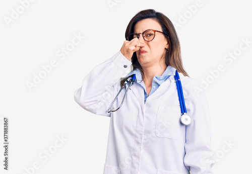 Young beautiful woman wearing doctor stethoscope and glasses smelling something stinky and disgusting  intolerable smell  holding breath with fingers on nose. bad smell