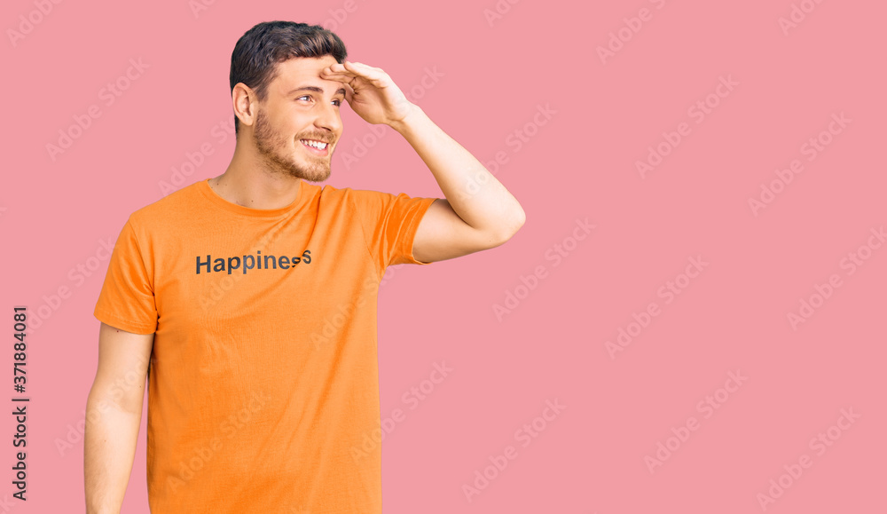 Handsome young man with bear wearing tshirt with happiness word message very happy and smiling looking far away with hand over head. searching concept. Handsome young man with bear wearing tshirt with happiness word message very happy and smiling looking far away with hand over head. searching concept.