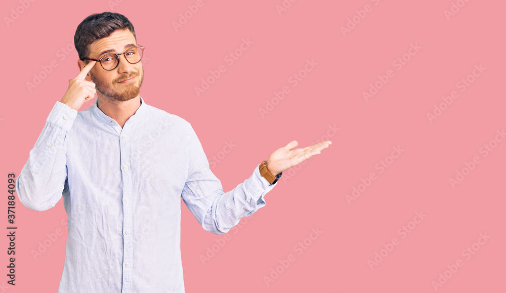 Handsome young man with bear wearing elegant business shirt and glasses confused and annoyed with open palm showing copy space and pointing finger to forehead. think about it. Handsome young man with bear wearing elegant business shirt and glasses confused and annoyed with open palm showing copy space and pointing finger to forehead. think about it.