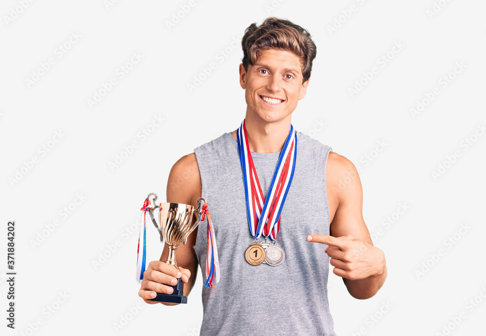 Young handsome man holding champion trophy wearing medals smiling happy pointing with hand and finger Young handsome man holding champion trophy wearing medals smiling happy pointing with hand and finger