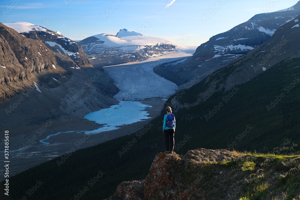 Woman trekker with backpack standing on steep cliff above the melting glacier and glacial lake in Columbia Icefield. Jasper National Park. Alberta. Canada Woman trekker with backpack standing on steep cliff above the melting glacier and glacial lake in Columbia Icefield. Jasper National Park. Alberta. Canada
