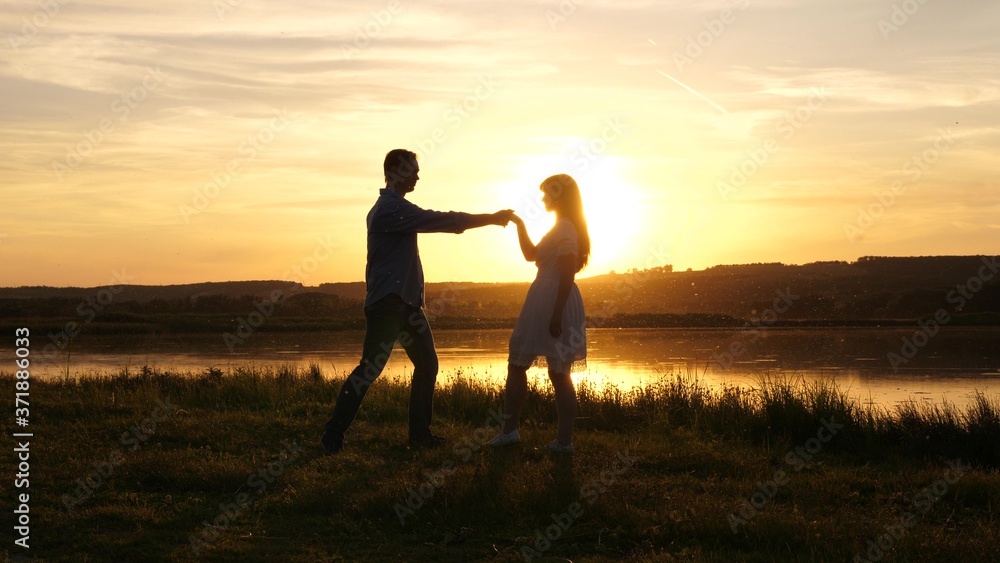 Young free couple dancing at sunset on beach. Happy guy and girl waltz in evening in a summer park. Enamored man and woman dance in the bright rays of the sun on background of the lake.