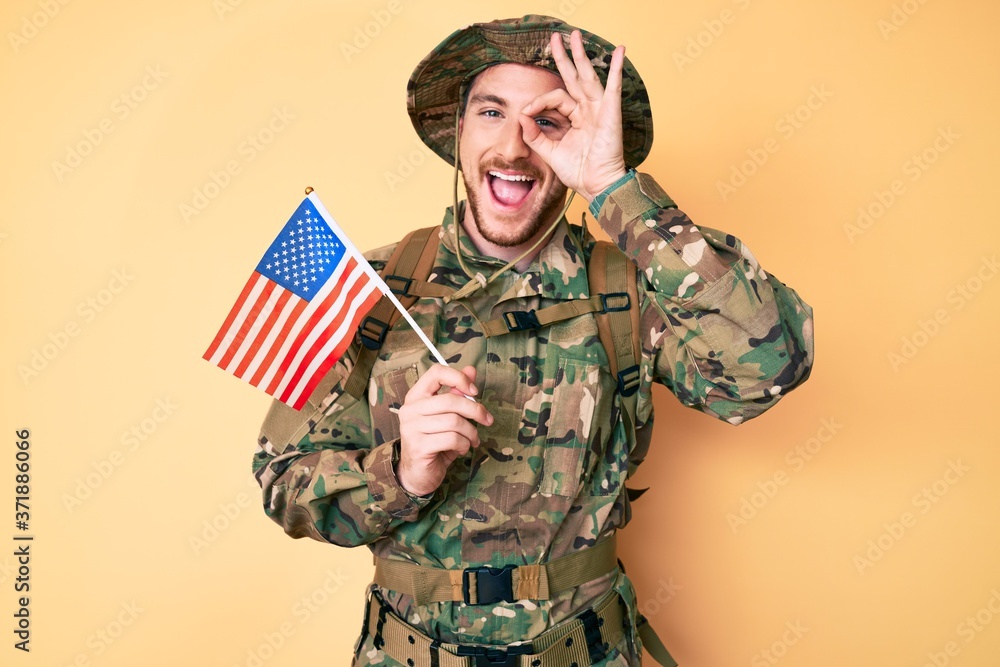 Young caucasian man wearing camouflage army uniform holding usa flag smiling happy doing ok sign with hand on eye looking through fingers