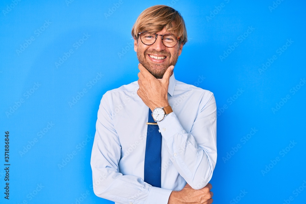 Young blond man wearing business clothes and glasses looking confident at the camera smiling with crossed arms and hand raised on chin. thinking positive. Young blond man wearing business clothes and glasses looking confident at the camera smiling with crossed arms and hand raised on chin. thinking positive.