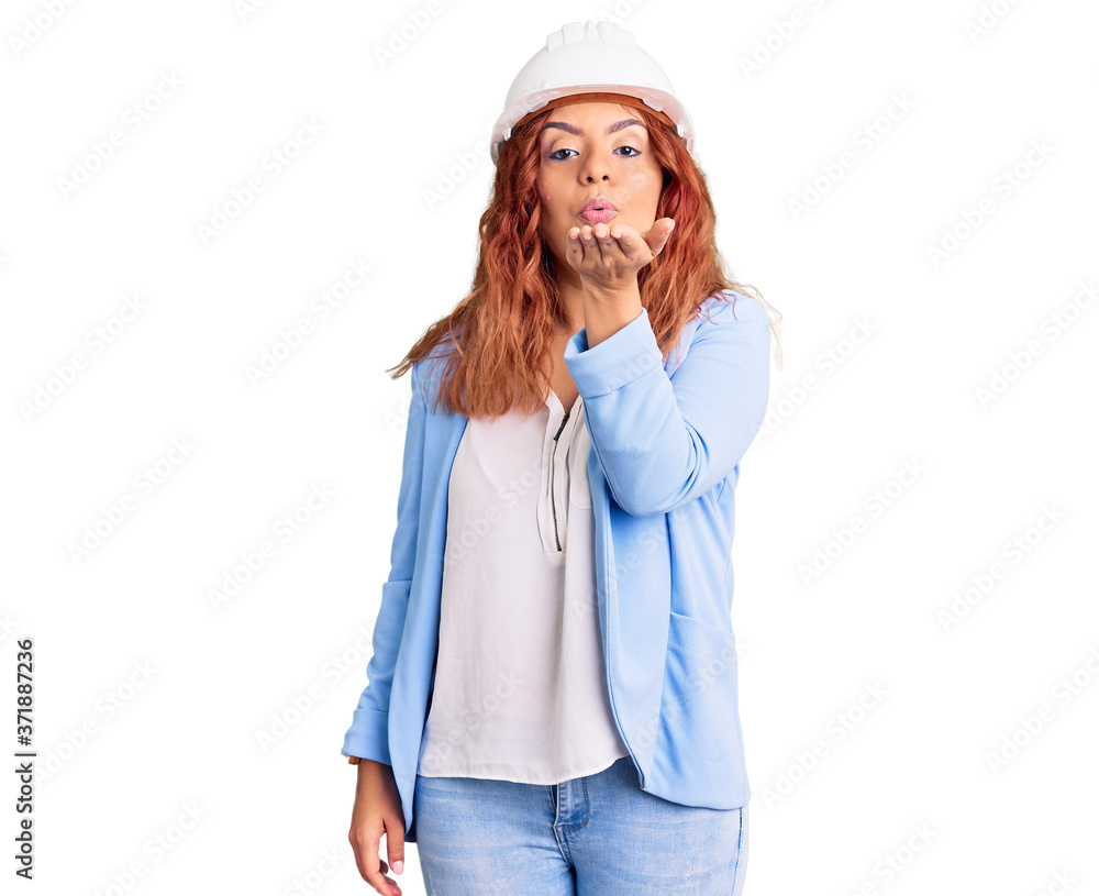Young latin woman wearing architect hardhat looking at the camera blowing a kiss with hand on air being lovely and sexy. love expression.