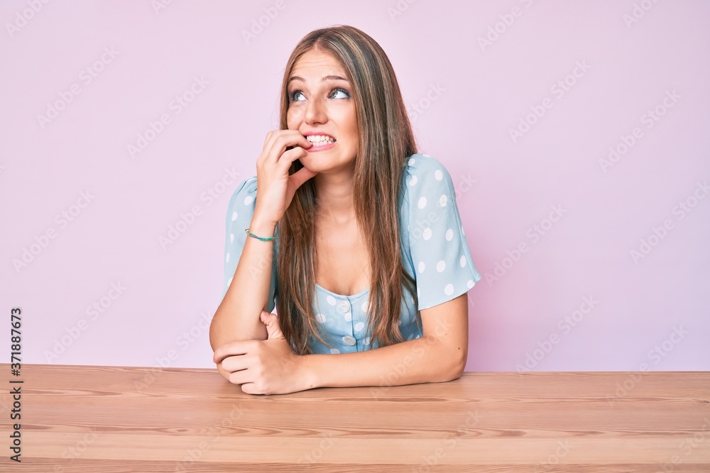 Young blonde girl wearing casual clothes sitting on the table looking stressed and nervous with hands on mouth biting nails. anxiety problem. Young blonde girl wearing casual clothes sitting on the table looking stressed and nervous with hands on mouth biting nails. anxiety problem.