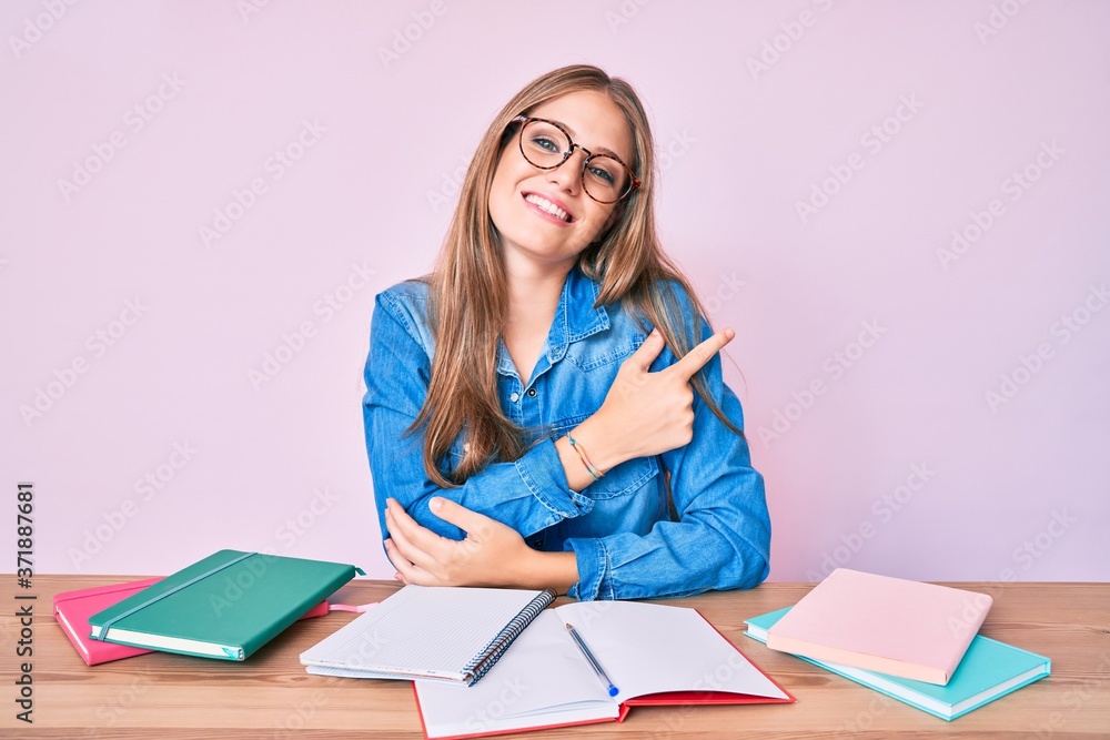 Young blonde girl studying sitting on the table smiling cheerful pointing with hand and finger up to the side Young blonde girl studying sitting on the table smiling cheerful pointing with hand and finger up to the side