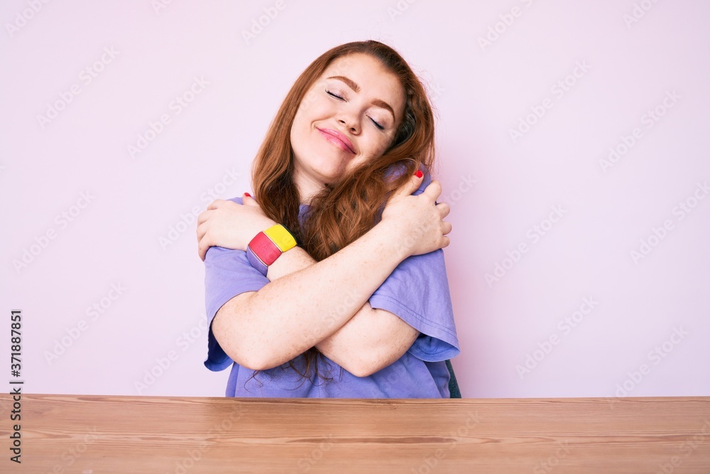 Young redhead woman wearing casual clothes sitting on the table hugging oneself happy and positive, smiling confident. self love and self care Young redhead woman wearing casual clothes sitting on the table hugging oneself happy and positive, smiling confident. self love and self care
