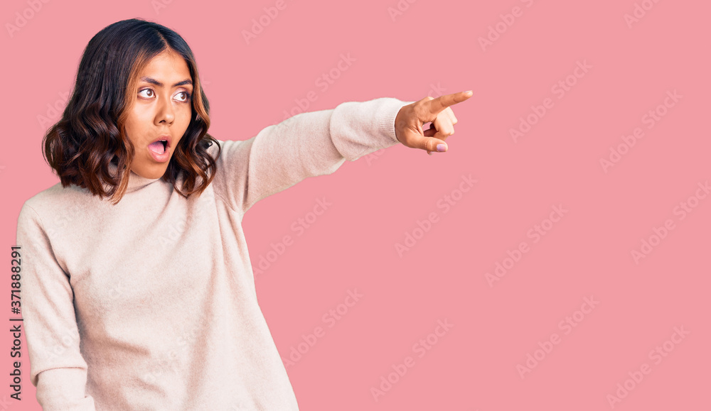 Young beautiful mixed race woman wearing winter turtleneck sweater pointing with finger surprised ahead, open mouth amazed expression, something on the front Young beautiful mixed race woman wearing winter turtleneck sweater pointing with finger surprised ahead, open mouth amazed expression, something on the front