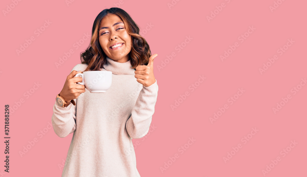 Young beautiful mixed race woman holding a cup of coffee smiling happy and positive, thumb up doing excellent and approval sign Young beautiful mixed race woman holding a cup of coffee smiling happy and positive, thumb up doing excellent and approval sign