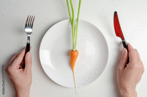 Woman holding fork and kinfe, ready to eat. Diet concept. One carrot on the plate on whte table. photo