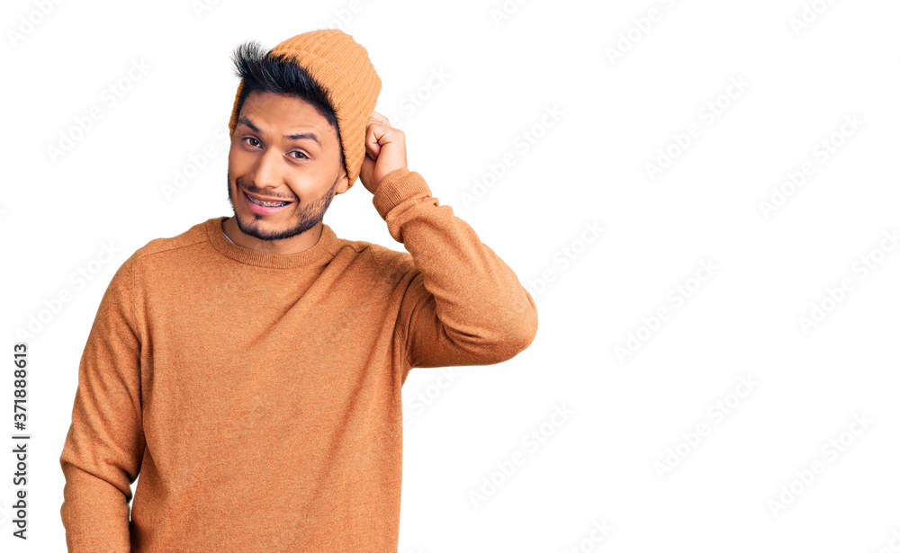 Handsome latin american young man wearing winter sweater and wool hat confuse and wonder about question. uncertain with doubt, thinking with hand on head. pensive concept. Handsome latin american young man wearing winter sweater and wool hat confuse and wonder about question. uncertain with doubt, thinking with hand on head. pensive concept.