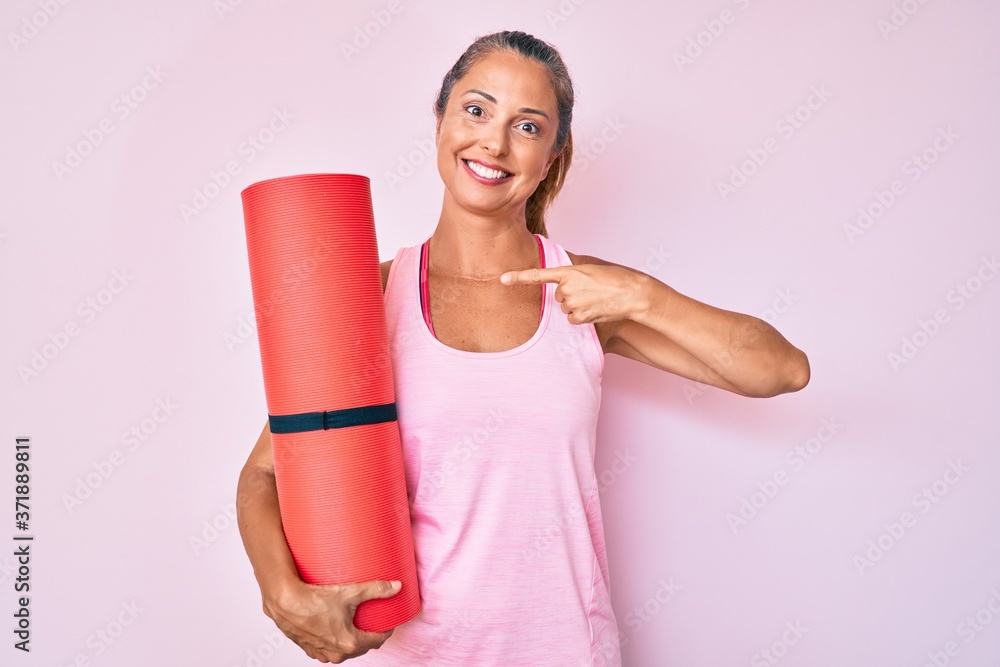 Middle age hispanic woman holding yoga mat pointing finger to one self smiling happy and proud