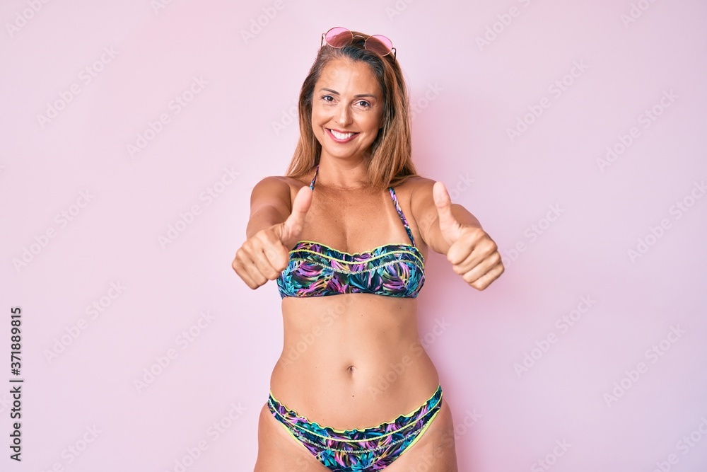 Naklejka premium Middle age hispanic woman wearing bikini approving doing positive gesture with hand, thumbs up smiling and happy for success. winner gesture.