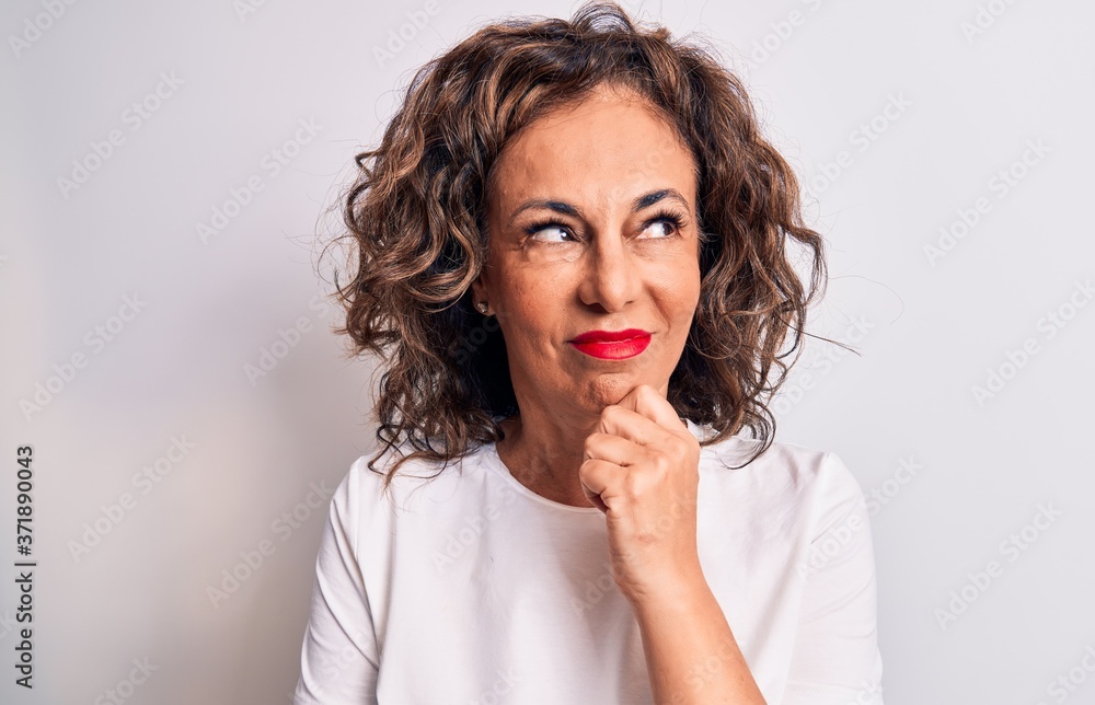 Middle age beautiful brunette woman wearing casual t-shirt standing over white background thinking concentrated about doubt with finger on chin and looking up wondering Middle age beautiful brunette woman wearing casual t-shirt standing over white background thinking concentrated about doubt with finger on chin and looking up wondering