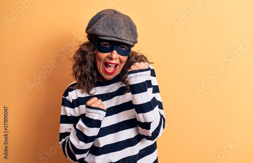 Middle age brunette burglar woman wearing mask and cap standing over yellow background celebrating surprised and amazed for success with arms raised and eyes closed