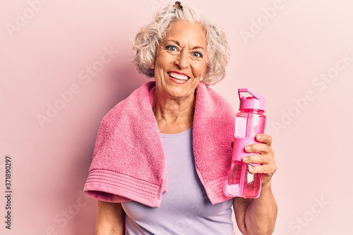 Senior grey-haired woman wearing sportswear and towel drinking bottle of water looking positive and happy standing and smiling with a confident smile showing teeth
