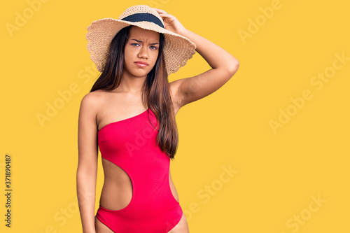 Young beautiful latin girl wearing swimwear and summer hat confuse and wonder about question. uncertain with doubt, thinking with hand on head. pensive concept.
