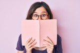 Young beautiful woman wearing glasses reading book afraid and shocked with surprise and amazed expression, fear and excited face.