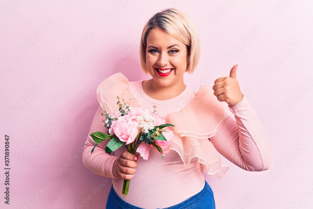 Beautiful blonde plus size woman holding bouquet of pink flowers over isolated background smiling happy and positive, thumb up doing excellent and approval sign
