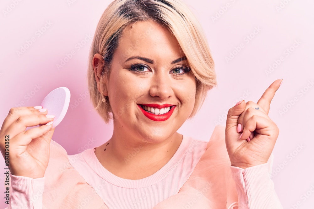 Young beautiful blonde plus size woman applying makeup using sponge over pink background smiling happy pointing with hand and finger to the side Young beautiful blonde plus size woman applying makeup using sponge over pink background smiling happy pointing with hand and finger to the side