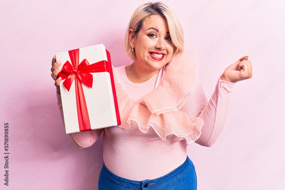 Young beautiful blonde plus size woman holding birthday gift over isolated pink background screaming proud, celebrating victory and success very excited with raised arm Young beautiful blonde plus size woman holding birthday gift over isolated pink background screaming proud, celebrating victory and success very excited with raised arm