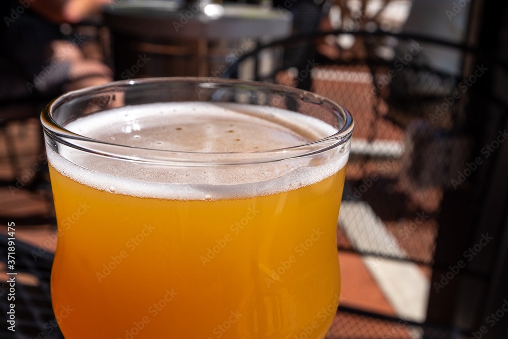 A clear glass of amber beer on a glass table at a micro brewery. The background has red tiles, metal tables and chairs. The glass of draft beer is full with a small white head on the top of the liquid A clear glass of amber beer on a glass table at a micro brewery. The background has red tiles, metal tables and chairs. The glass of draft beer is full with a small white head on the top of the liquid