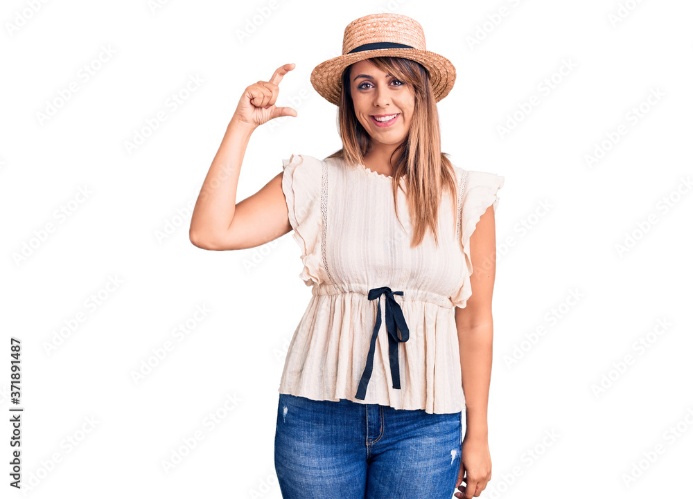 Young beautiful woman wearing summer hat and t-shirt smiling and confident gesturing with hand doing small size sign with fingers looking and the camera. measure concept. Young beautiful woman wearing summer hat and t-shirt smiling and confident gesturing with hand doing small size sign with fingers looking and the camera. measure concept.