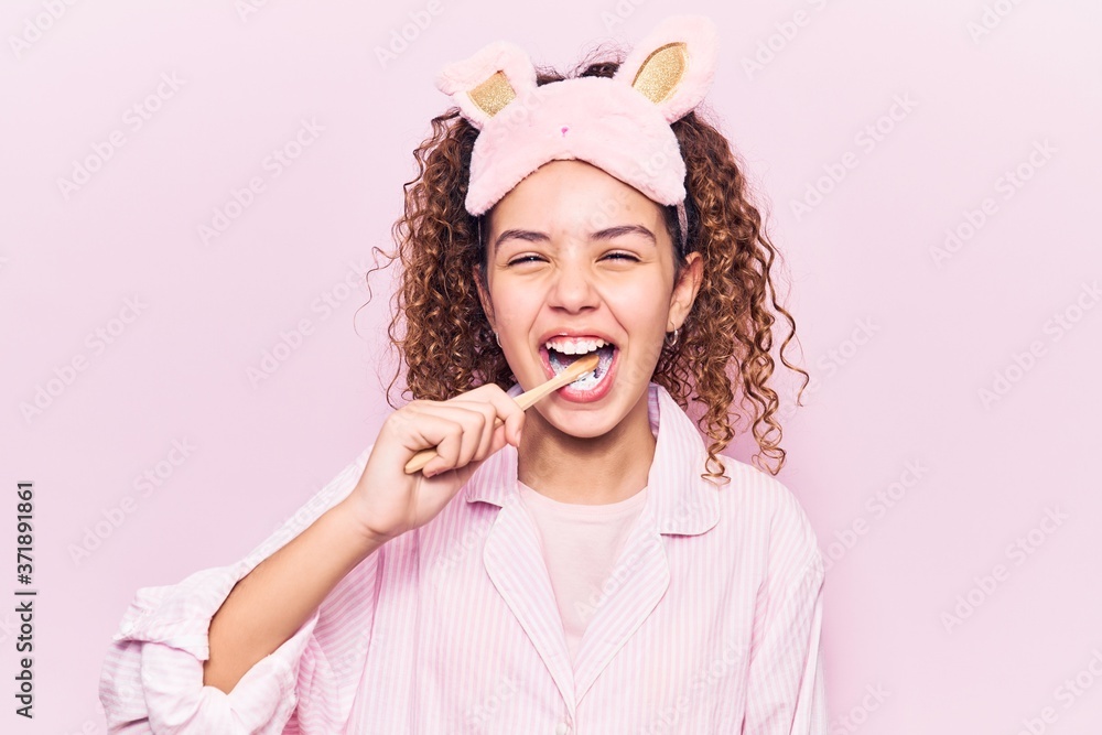 Beautiful latin teenager wearing pajama and sleep mask smiling happy. Standing with smile on face using toothbrush over isolated pink background Beautiful latin teenager wearing pajama and sleep mask smiling happy. Standing with smile on face using toothbrush over isolated pink background