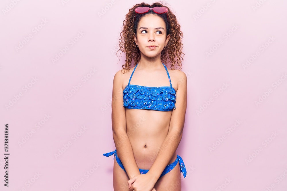 Naklejka premium Beautiful kid girl with curly hair wearing bikini and sunglasses smiling looking to the side and staring away thinking.