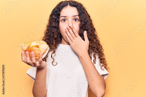 Beautiful kid girl with curly hair holding potato chip covering mouth with hand, shocked and afraid for mistake. surprised expression