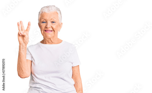 Senior beautiful woman with blue eyes and grey hair wearing casual white tshirt showing and pointing up with fingers number three while smiling confident and happy.