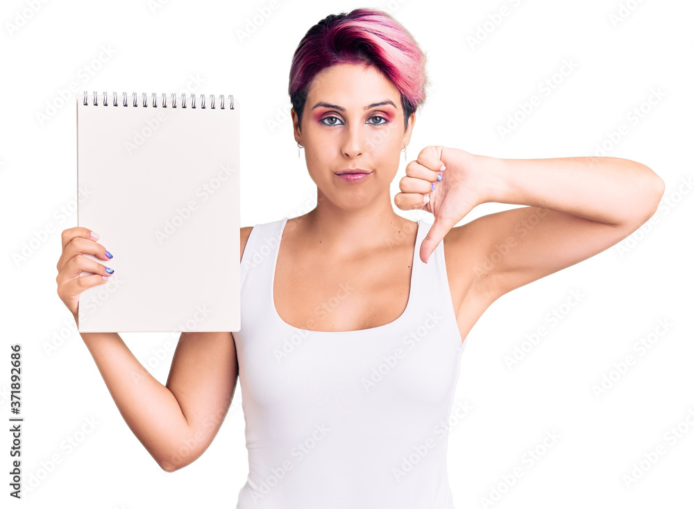 Young beautiful woman with pink hair holding notebook with angry face, negative sign showing dislike with thumbs down, rejection concept Young beautiful woman with pink hair holding notebook with angry face, negative sign showing dislike with thumbs down, rejection concept
