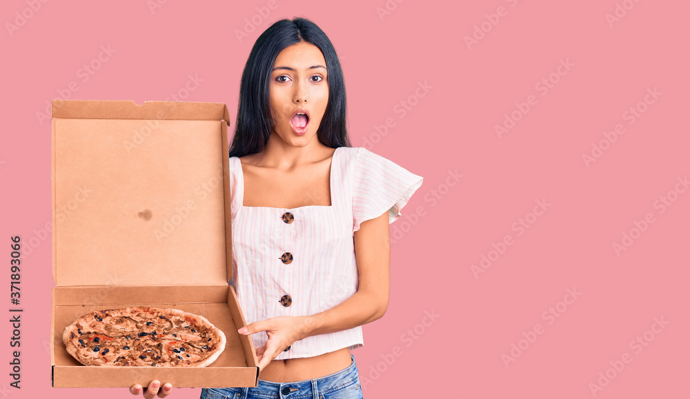 Young beautiful latin girl holding delivery pizza box scared and amazed with open mouth for surprise, disbelief face Young beautiful latin girl holding delivery pizza box scared and amazed with open mouth for surprise, disbelief face
