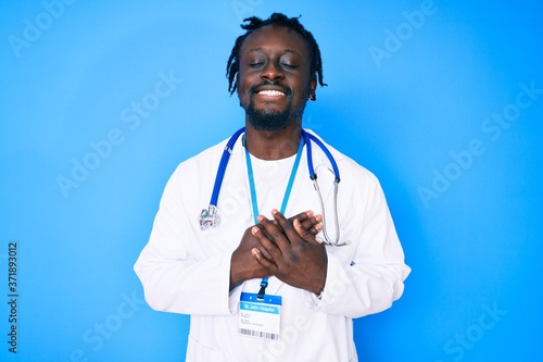 Young african american man with braids wearing doctor stethoscope and id pass smiling with hands on chest with closed eyes and grateful gesture on face. health concept. photo